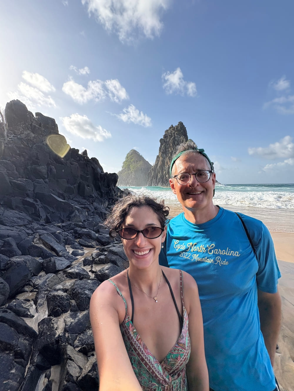 Father and daughter taking a selfie on a rocky beach with the Morro Dois Irmãos rock formations and the ocean in the background.