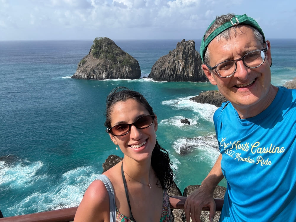 Father and daughter smiling for a selfie at a viewpoint overlooking the turquoise ocean and the iconic Morro Dois Irmãos rock formations.