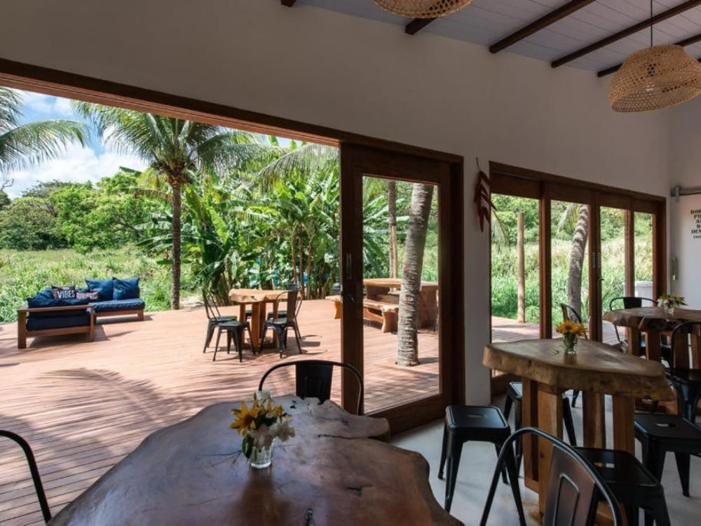 Open-air common area of a hostel with a rustic wooden table and wide doors showing lush vegetation in Fernando de Noronha.