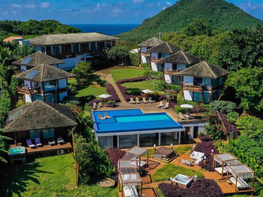 Aerial view of Pousada Morena featuring dark-roofed cabins, a central blue swimming pool, and hot tubs surrounded by lush forests in Fernando de Noronha.