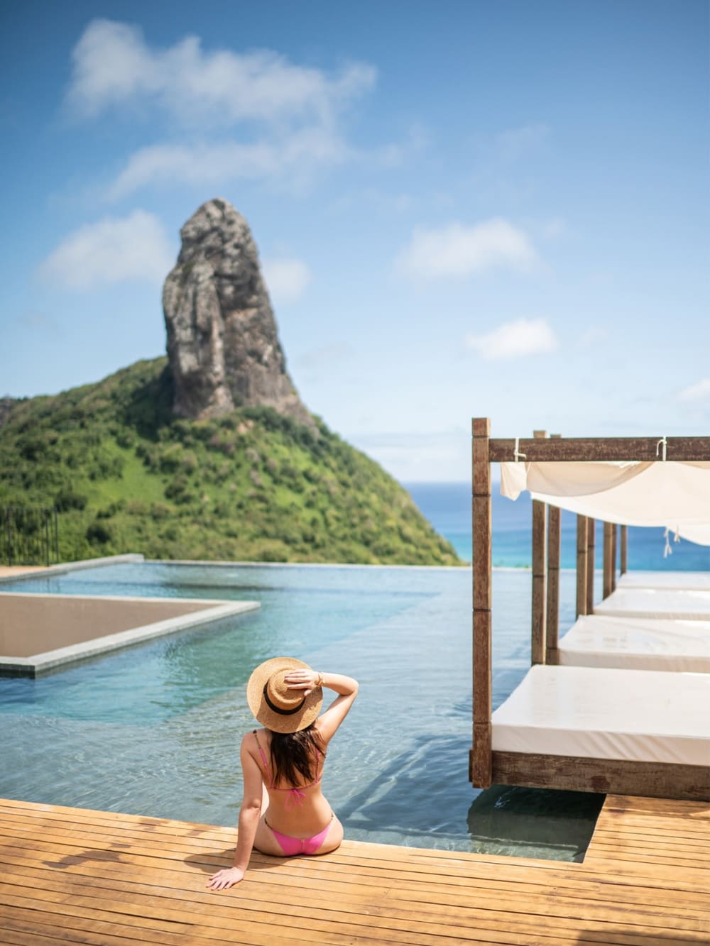Woman wearing a straw hat relaxing on a wooden deck by an infinity pool with a stunning view of Morro do Pico at a luxury hotel in Fernando de Noronha, Brazil.