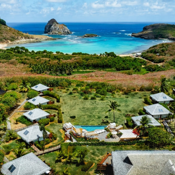 Aerial view of a luxury eco-resort featuring thatched-roof bungalows, green lawns, and a central pool area near the pristine coastline of Fernando de Noronha.