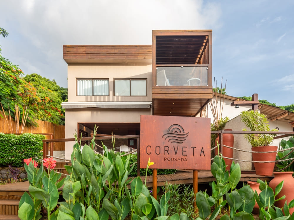 Modern exterior of Pousada Corveta with a metal sign, wooden walkways, and tropical plants, highlighting where to stay in Fernando de Noronha.