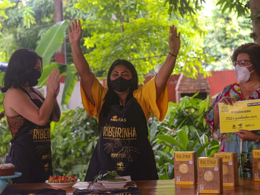 Uma mulher usando um avental preto com a marca FAS Ribeirinha levanta as mãos de alegria, ao lado de outras pessoas celebrando o programa da Fundação Amazônia Sustentável.