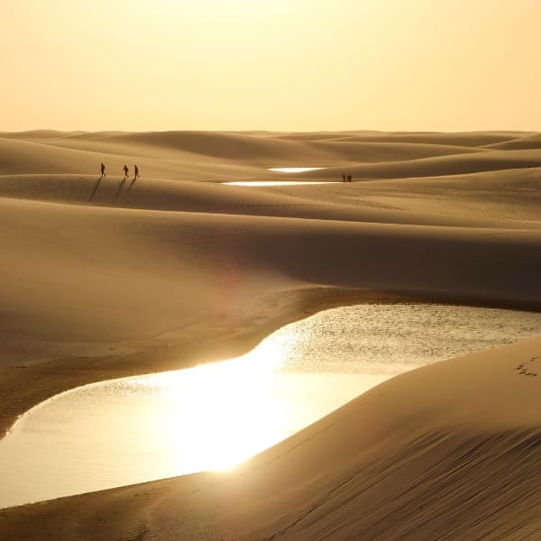 Silhouettes of people walking along vast sand dunes near small rainwater lagoons during a golden sunset in Lencois Maranhenses.