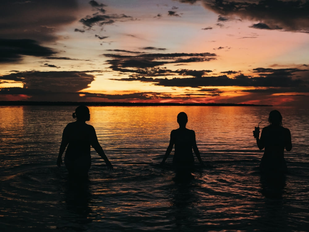 Silhouettes of three people standing waist-deep in the waters of the Amazon at sunset.