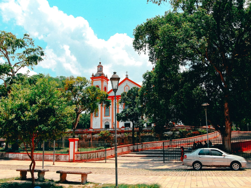 White and orange historic church with a curved ramp surrounded by green trees in a quiet Manaus city plaza. 