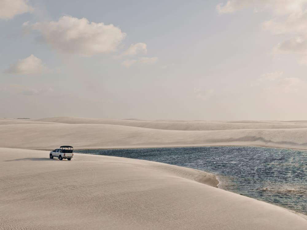 A white 4x4 tour vehicle driving across vast white sand dunes next to a bright blue lagoon.
