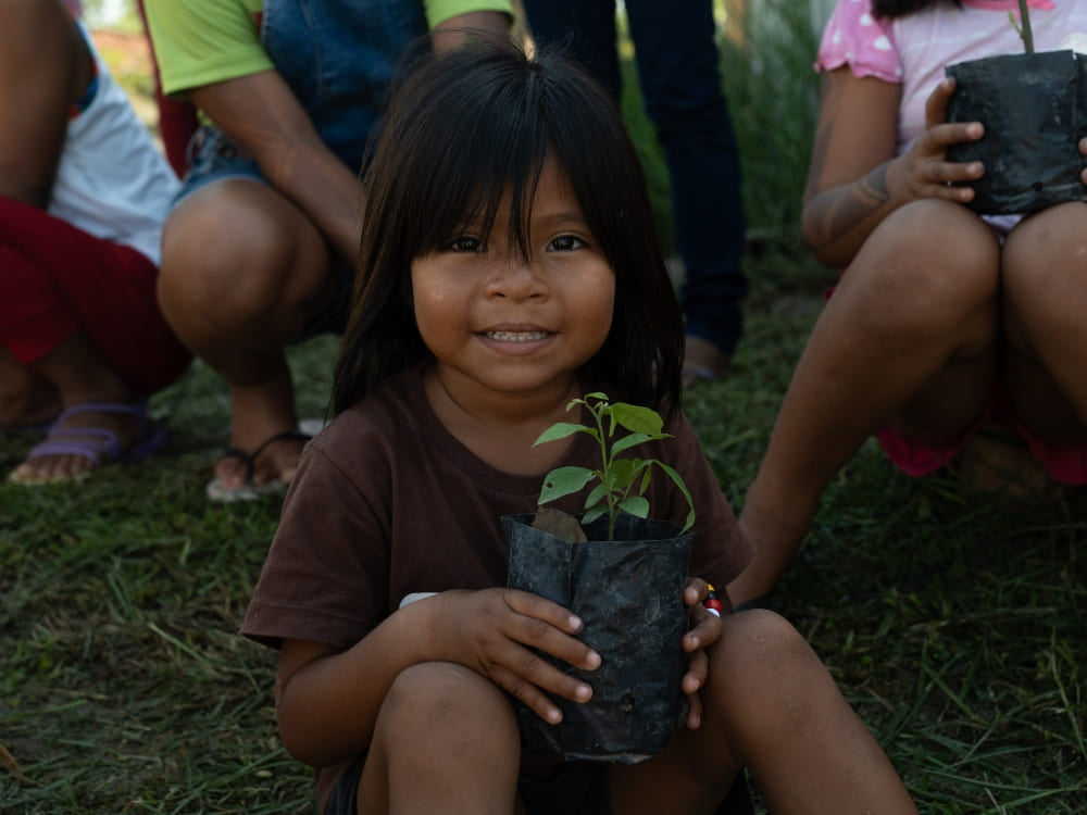 A smiling child from an Amazonian riverside community sitting on the grass and holding a small potted plant.