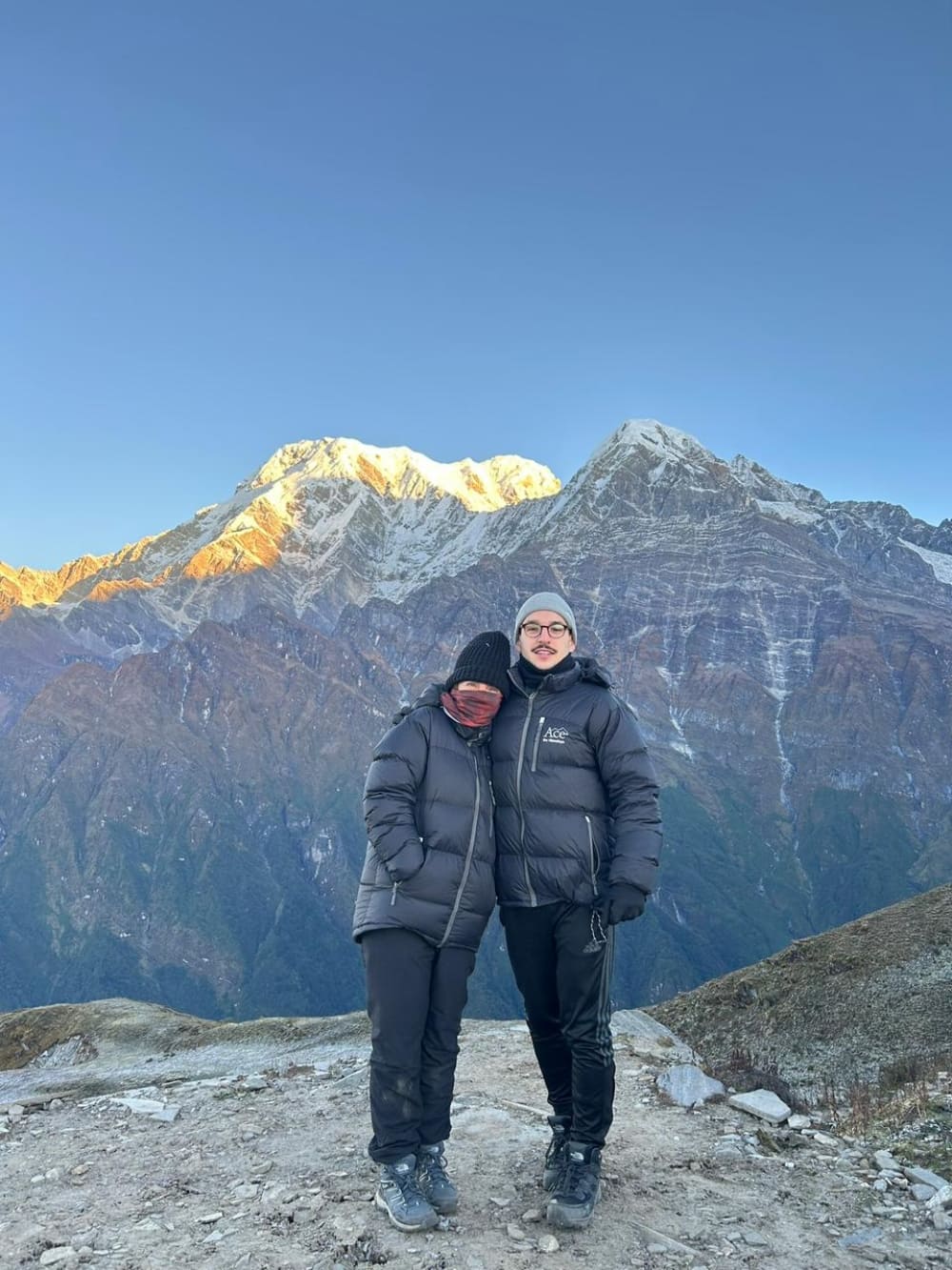 Mother and son Mila and Lorenzo wearing heavy winter gear and embracing in front of the snowy Himalayan peaks after an early morning climb.