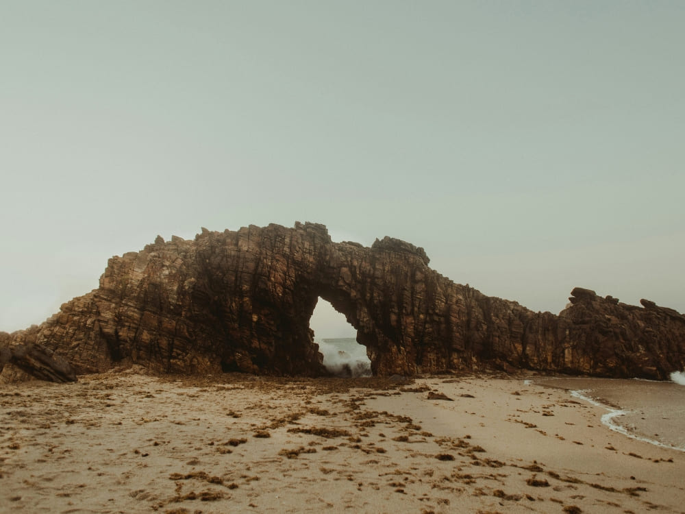 A large, rugged rock formation featuring a natural arch on a sandy beach beside the ocean, known as Pedra Furada in Jericoacoara. 