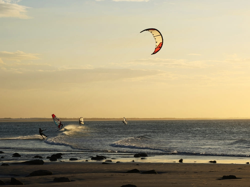 Silhouettes of people kitesurfing and windsurfing on ocean waves near a sandy beach during a golden sunset.