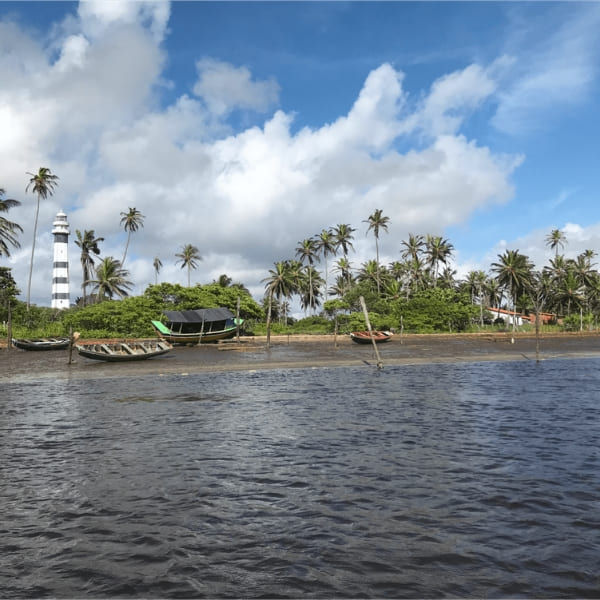 Waterfront view of Mandacaru community, featuring a black and white striped lighthouse, fishing boats on the shore, and coconut trees under a cloudy sky.