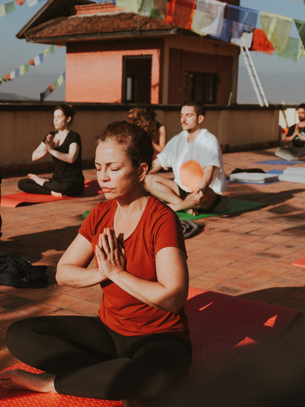 Mila Ricetti and Lorenzo practicing yoga and meditating with a group on a sunlit rooftop decorated with prayer flags during a retreat in Nepal.
