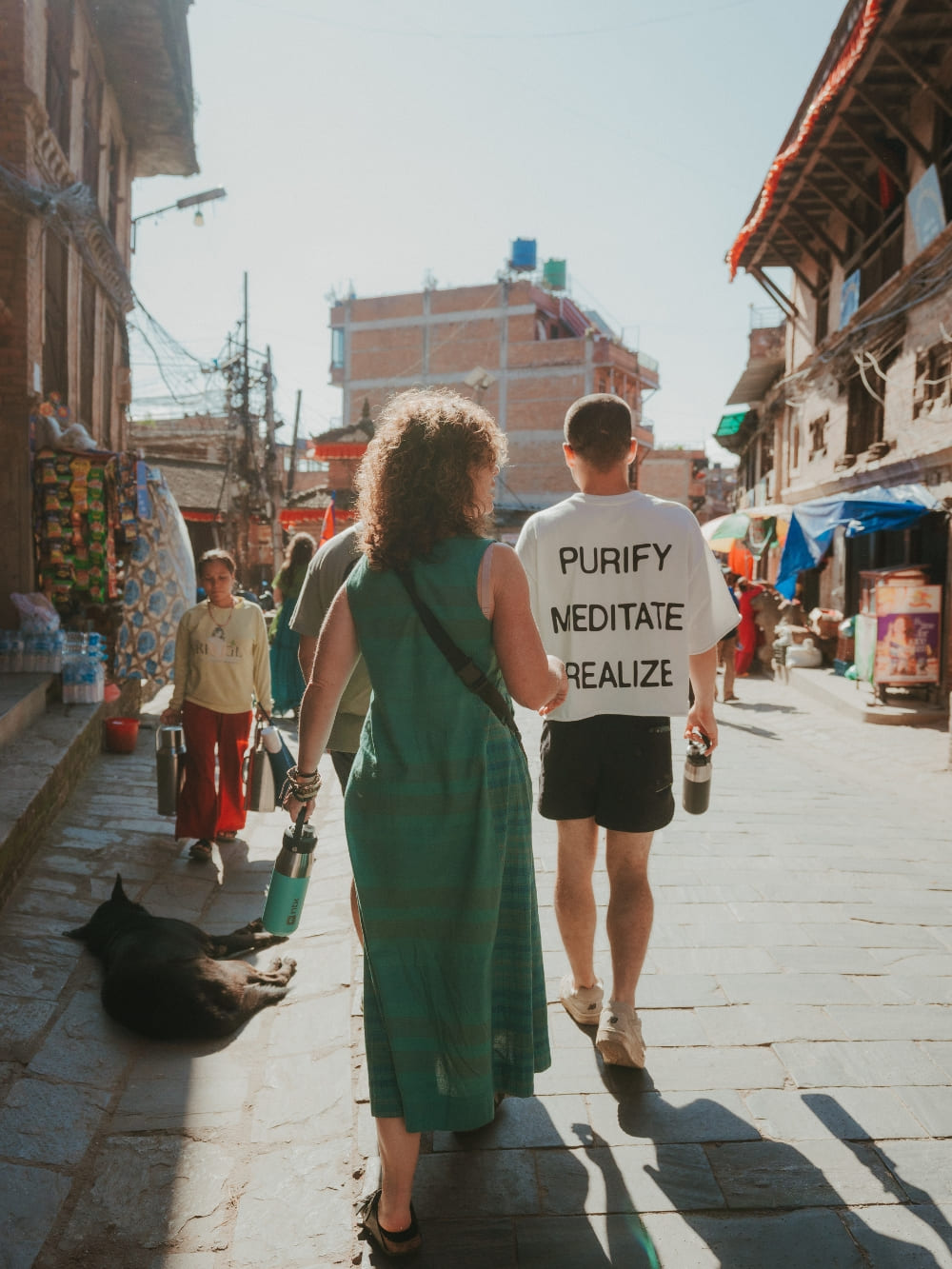 Mila and Lorenzo walking side by side down a sunny street in Nepal, with Lorenzo wearing a t-shirt that says purify, meditate, realize.